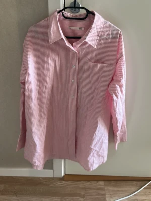 Rosa och vit randig skjorta - Säljer en klassisk rosa och vit randig skjorta med lång ärm och knappar framtill. Skjortan har en bröstficka och avslappnad passform, perfekt för en chill och stilren look. Tillverkad i bomull för en skön känsla hela dagen.