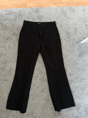 Svarta kostymbyxor från Gina Tricot - Snygga svarta kostymbyxor från Gina Tricot med klassisk rak passform och pressveck. Byxorna har hällor för skärp och sidofickor. Perfekta för en stilren look och lätta att matcha med olika toppar.