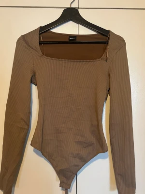 Ribbad brun långärmad body  - Säljer en ribbad body från Gina Tricot i brun nyans. Bodyn har lång ärm, fyrkantig urringning och är tillverkad i stretchigt material som sitter snyggt på kroppen. Perfekt att styla med jeans eller kjol för en trendig look.
