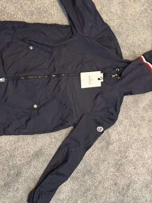 Mörkblå Moncler vindjacka med huva - Snygg mörkblå vindjacka från Moncler med huva och dragkedja framtill. Jackan har två fickor med tryckknappar, Moncler-logga på ärmen och en rödvit rand på huvan. Tillverkad i lätt och smidigt material, perfekt för blåsiga dagar.