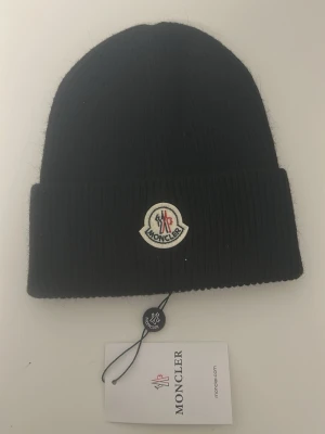 Svart Moncler mössa med logotyp - Snygg svart ribbstickad mössa från Moncler med uppvikt kant och klassisk Moncler-logga framtill. Perfekt accessoar för att hålla värmen och ge din outfit en exklusiv touch.