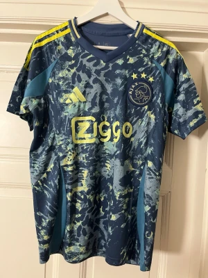 Ajax fotbollströja blå/gul Adidas - Ajax fotbollströja i blått med gula detaljer och mönster. Korta ärmar, rund hals och klubbmärke på bröstet. Adidas-logga och Ziggo-sponsor i gult. Perfekt för fotbollsträning eller match.