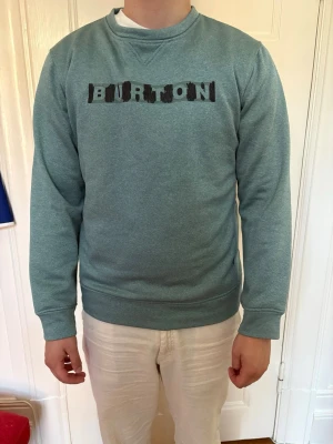 Grön sweatshirt från Burton - Snygg blå/grön sweatshirt från Burton med svart tryck på bröstet. Tröjan har rund halsringning, långa ärmar och ribbade muddar vid ärmslut och nederkant. Färg: lite åt det grön turkosa hållet, slitet tryck fram.
