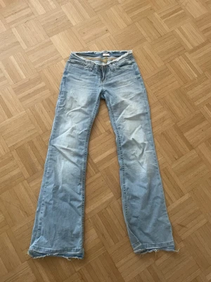 Ljusblå bootcut jeans från Nelly - Säljer ett par ljusblå bootcut jeans från Nelly med råa, fransiga kanter i midjan och vid bensluten. Jeansen har klassisk femficksdesign och är tillverkade i denim med en snygg tvättad look. Perfekta för dig som gillar en avslappnad och trendig stil.