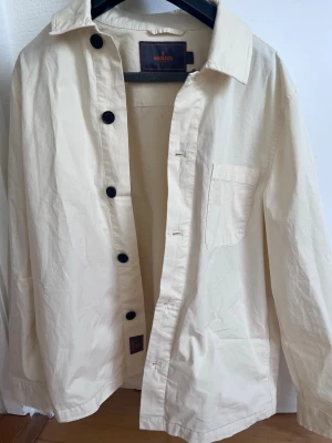 Kritvit overshirt från Morris - Stilren overshirt från Morris i kritvit bomull. Klassisk krage, svarta knappar och en bröstficka ger en clean look. Perfekt lager-på-lager-plagg för dig som gillar minimalistisk stil och vill ha något lätt och snyggt till vardags.