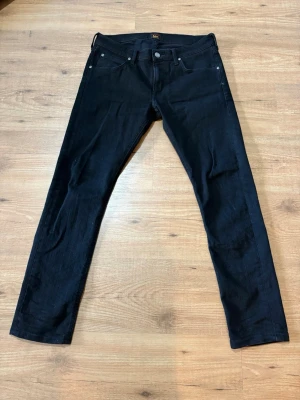 Svarta jeans från Lee - Säljer ett par svarta jeans från Lee med klassisk femficksmodell och raka ben. Jeansen har silverfärgade nitar och knapp, samt Lee-logga på bakfickan och bakre linningen. Materialet är denim i bomull med en snygg svart tvätt.
