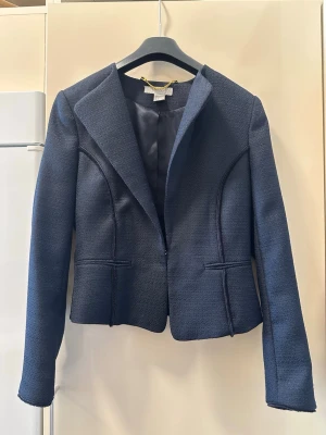 Mörkblå kavaj från H&M, storlek 36 - Stilren mörkblå kavaj från H&M i storlek 36. Kavajen har markerade axlar, figurnära passform och är tillverkad i ett strukturerat tyg med snyggt foder. Klassisk krage och två fickor framtill. Perfekt för en dressad look. Kavajen har lite defekter som kan ses på sista bilden💙