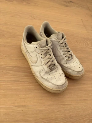Nike Air Force 1 vita sneakers - Klassiska vita Nike Air Force 1 sneakers med lågt skaft och ikonisk swoosh på sidan. Skorna är i läder med perforerade detaljer på tån och har en tjock sula med AIR-logga bak. Perfekta för en clean och tidlös streetstil.