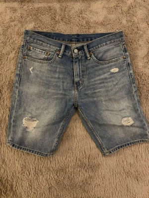 Levi's blå jeansshorts med slitningar - Säljer ett par klassiska blå jeansshorts från Levi's med snygga slitningar. Shortsen har normal passform, knappgylf och är tillverkade i bomull. Perfekta för sommaren och ger en avslappnad streetstyle-look. Lite sliten på lappen (syns på bild 3)
