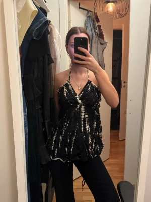 Tie-dye halterneck topp med volanger – svart/vit | Strl S/M  - Snygg och unik halterneck topp i tie-dye mönster i svart och vitt. Djup v-ringning fram, öppen rygg med tunna snörningar längs ryggen och volangkant nedtill som ger ett feminint och bohemiskt intryck. Resår under bysten framhäver figuren. Passar både S och M då snörningen i ryggen är justerbar.  Märke okänt, använd men i gott skick. 