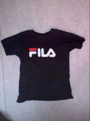 Svart FILA t-shirt med logga - Säljer en svart t-shirt från FILA med stor vit och röd logga på bröstet. Klassisk passform och korta ärmar, perfekt till jeans eller shorts. Tillverkad i mjuk bomull för skön känsla hela dagen.
