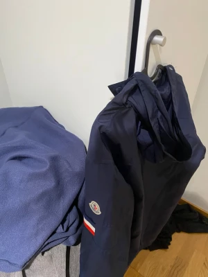 Mörkblå Moncler vindjacka med huva - Snygg mörkblå vindjacka från Moncler med huva och tryckknappar framtill. Jackan har Moncler-logga på ärmen och detaljer i rött, vitt och blått på båda ärmarna. Materialet är lätt och vattenavvisande, perfekt för blåsiga dagar.