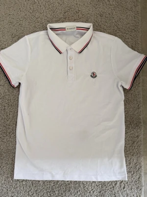 Vit Moncler pikétröja med röd detalj - Klassisk vit pikétröja från Moncler med korta ärmar och krage med röd och marinblå rand. Tröjan har tre knappar framtill och Moncler-logga broderad på bröstet. Perfekt till jeans eller shorts för en clean och sportig look.