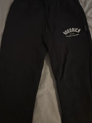 Svarta mjukisbyxor från Hoodrich - Säljer ett par svarta mjukisbyxor från Hoodrich med tryckt logga på vänster ben. Byxorna har dragsko i midjan och resår, samt sidofickor. Perfekta för chill dagar och streetwear-stil.