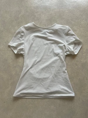Vit basic t-shirt i bomull - En enkel vit t-shirt i mjuk bomull med rund hals och korta ärmar. Modellen har en klassisk passform och är perfekt för en clean och stilren look. Inga synliga tryck eller detaljer.