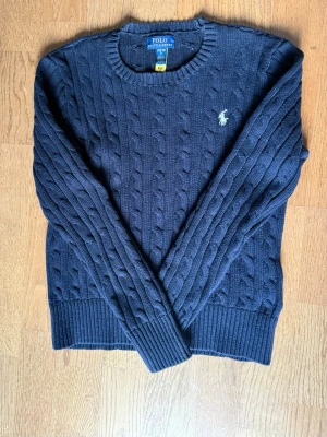 Mörkblå tröja Polo Ralph Lauren - Mörkblå kabelstickad tröja från Polo Ralph Lauren med rund halsringning och vit logga. Väldigt fint skick, men skulle säga att den är lite liten i storlek. Ålder 12-14 står det på storleken. 