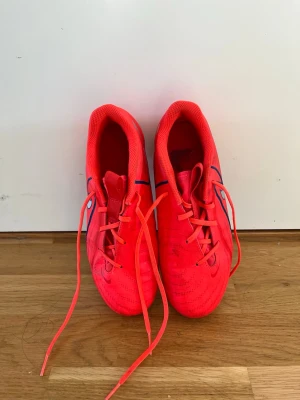 Nike röda fotbollsskor med dobbar - Säljer ett par röda Nike fotbollsskor med svarta detaljer och klassisk snörning. Skorna har fasta dobbar för spel på gräs och är tillverkade i syntetmaterial som ger bra passform och känsla på planen. Perfekta för dig som vill sticka ut på matchen!