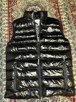 Svart glansig dunväst från Moncler - Snygg svart dunväst från Moncler med glansig finish och klassisk logga på bröstet. Västen har hög krage, två dragkedjefickor och tydlig Moncler-detalj på dragkedjan. Perfekt för lager-på-lager och streetstyle.