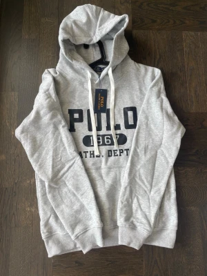 Grå hoodie från Polo Ralph Lauren - Helt ny och oanvänd grå hoodie från Polo Ralph Lauren med stort tryck framtill. Storlek M men liten i storleken, passar även S. Skön och stilren hoodie med huva och vita dragsnören