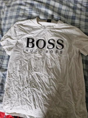 Vit Hugo Boss t-shirt med logga - Säljer en vit t-shirt från Hugo Boss med stor svart logga på bröstet. Klassisk rund halsringning och korta ärmar. Tillverkad i mjuk bomull för skön passform. Perfekt för dig som gillar stilrena och tidlösa plagg med tydlig branding. Passar mig inte längre. Bra o tvätta så blir den fin o len