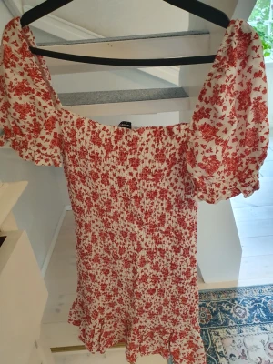 Blommig kortklänning Stradivarius - Supersöt kortklänning från Stradivarius med rödblommigt mönster på vit botten. Klänningen har puffiga korta ärmar, smockad överdel och volangkant nertill. Perfekt för sommarens alla tillfällen och ger en romantisk vibe.