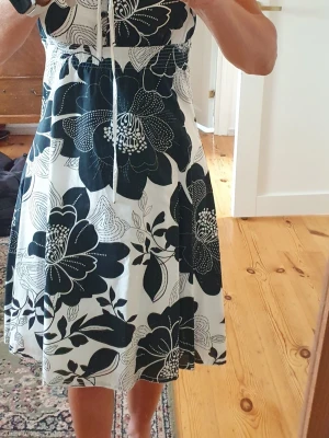 Svartvit blommig sommarklänning - Snygg svartvit klänning med stora blommor och smala axelband. Klänningen har snörning framtill och är i en luftig, lätt modell som passar perfekt till varma dagar. Midjan är markerad med resår och kjolen har en ledig passform.