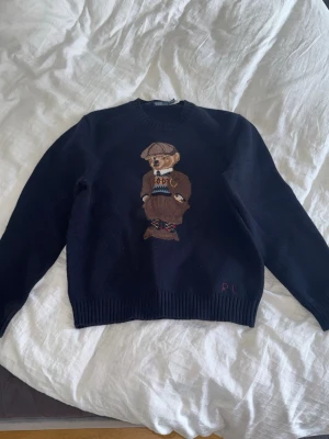 Mörkblå Polo Bear tröja Ralph Lauren - Mörkblå stickad tröja från Polo Ralph Lauren med ikoniska Polo Bear-motivet framtill. Rund halsringning och ribbade muddar. Perfekt för dig som gillar klassisk och lekfull stil. XL men passar L