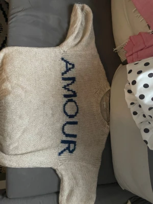 Beige stickad tröja med blå text - Stickad beige tröja från Design by Si med blå texten 'AMOUR' på framsidan. Tröjan har rund halsringning och är gjord i ett mjukt, grovt garn som ger en avslappnad och mysig känsla. Perfekt för dig som gillar statement-plagg och vill ha något unikt i garderoben.