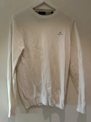 Vit stickad tröja från GANT - Snygg vit stickad tröja från GANT med diskret logga broderad på bröstet. Tröjan har rund halsringning, ribbade muddar och är tillverkad i ett mjukt, strukturerat material. Perfekt för en stilren och avslappnad look.