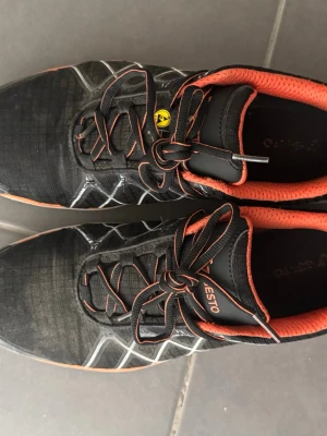Svarta skyddskor/arbetsskor från Vestro med orange detaljer - Snygga svarta sneakers från Vestro med orange innersula och kant, samt vita och orange detaljer på sidorna. Skorna har snörning med matchande orange snören och en vit sula med orange rand. Materialet är textil och syntet, perfekt för en sportig look.