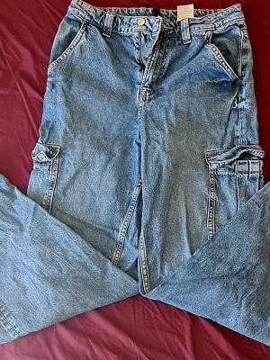 Baggy denim cargobyxor med fickor - Säljer ett par ljusblå baggy cargobyxor i denim med stora sidofickor och klassisk femficksdesign. Byxorna har bred passform och cool vintagekänsla, perfekta för en avslappnad streetstyle-look. Materialet är 100% bomullsdenim.