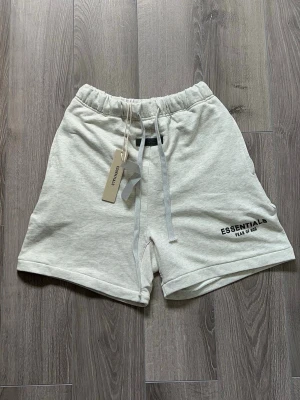 Essentials Fear of God shorts grå - Snygga ljusgrå shorts från Essentials Fear of God med elastisk midja och snörning. Mjuk bomullskänsla och logga tryckt på vänster ben. Perfekta för chill dagar och enkel streetwear-stil.