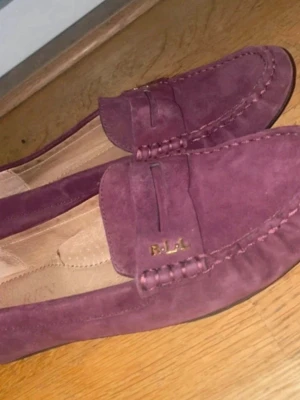 Vinröda loafers från Ralph Lauren - Säljer ett par vinröda loafers i mocka från Ralph Lauren med RL-logga i guld på sidan. Skorna har klassisk pennyloafers-design och gummisula med nabbad undersida för extra grepp. Perfekta för en stilren och avslappnad look. Bra skick! Storlek 37