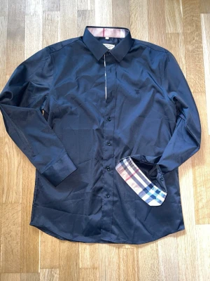 Svart skjorta från Burberry  - Snygg svart skjorta från Burberry med klassiskt rutigt mönster på insidan av manschetter och knappslå. Skjortan har lång ärm, knappar framtill och diskret broderad logga på bröstet. Tillverkad i mjuk bomull för en bekväm känsla. Sitter mer som M