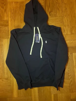 Mörkblå hoodie från Polo Ralph Lauren - Snygg mörkblå hoodie från Polo Ralph Lauren med vit broderad logga på bröstet och vita dragsnören i huvan. Klassisk känguruficka framtill och ribbade muddar vid ärmar och nederkant. Perfekt för en avslappnad och stilren look.