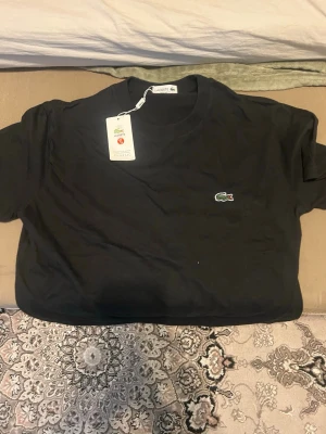 Svart t-shirt från Lacoste - Klassisk svart t-shirt från Lacoste med den ikoniska gröna krokodilloggan broderad på bröstet. T-shirten har rund hals och korta ärmar. Tillverkad i mjuk bomull för en skön känsla och enkel stil.