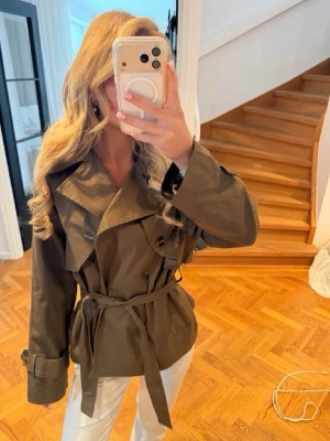 Olivgrön trenchcoat med knytskärp - Stilren olivgrön trenchcoat med dubbelknäppning, bred krage och knytskärp i midjan. Jackan har dekorativa axelklaffar och justerbara ärmslut med spännen. Perfekt för dig som vill ha en klassisk men trendig look.