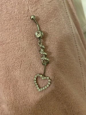 Silvrigt hjärtformat navel piercing  - Säljer ett silvrigt navel piercing med ett hängande hjärta dekorerat med gnistrande stenar. Navel piercingen har en böjd stav och flera små stenar i spiralform ovanför hjärtat. Perfekt för dig som gillar detaljer och bling.