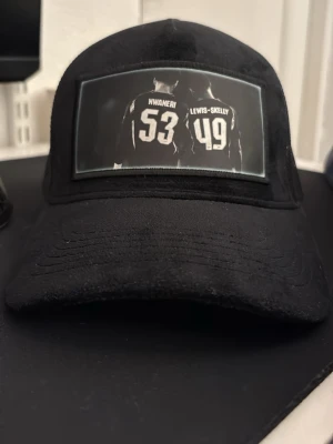 Svart truckerkeps med tryck - Säljer en svart truckerkeps med mesh baktill och böjd skärm. På framsidan finns ett tryck med två ryggar och siffrorna 53 och 49 samt namnen Nwaneri och Lewis-Skelly. Kepsen har justerbar storlek och är tillverkad i bomull och polyester.