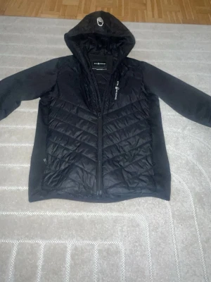 Svart jacka från sailracing (äkta) - Säljer en svart vindjacka från Sailracing med quiltad front och huva. Jackan har dragkedja, logotyp på bröstet och huvan samt släta ärmar. Materialet är syntet och jackan är perfekt för blåsiga dagar. Enkel och stilren design som passar till många outfits.