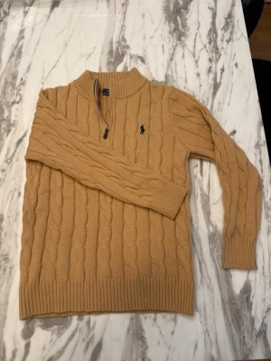 Beige kabelstickad tröja Polo Ralph Lauren - Kabelstickad beige tröja från Polo Ralph Lauren med halv dragkedja och hög krage. Tröjan har ett mörkblått broderat logotyp på bröstet och ribbade muddar vid ärmslut och nederkant. Perfekt för lager på lager och snygg till jeans.