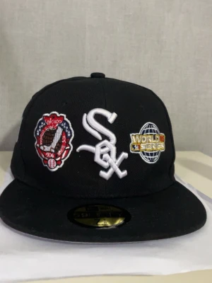 Svart White Sox World Series keps - Snygg svart keps från New Era med Chicago White Sox-logga framtill, broderade World Series-märken och texten 'Series Champions' på sidan. Klassisk flat brim och broderade detaljer i vitt, rött och guld. Perfekt för dig som gillar baseball och streetwear.