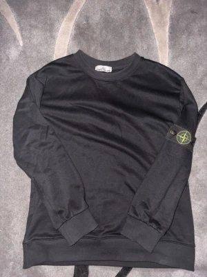 Svart långärmad topp från Stone Island - Svart långärmad topp från Stone Island med klassisk rund hals och ribbade muddar. På vänster ärm sitter det ikoniska Stone Island-märket. Enkel och stilren design som passar till mycket.