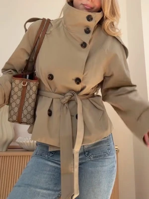 Bershka trenchcoat  - En väldigt snygg och populär trenchcoat från Bershka, den är i väldigt bra skick. Den är kort och beige, och har bruna knappar och ett bälte till. Väldigt stilren jacka som går att styla med det mesta. Säljs inte längre.