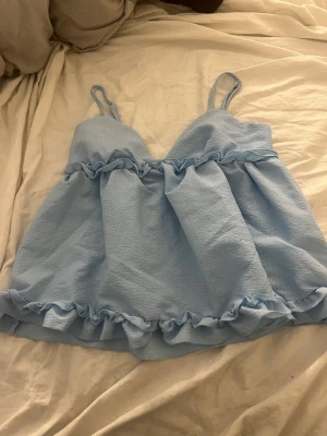 Ljusblå volangblus med smala band - Supersöt ljusblå blus med tunna axelband och volangdetaljer både upptill och nedtill. Blusen har en luftig och flowy passform. Perfekt för en somrig och chill look. Säljer då den är lite liten och inte helt min stil, aldrig använt!