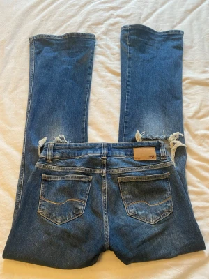 Blå bootcut jeans med slitningar 157 - Säljer ett par mörkblåa bootcut jeans från 157 med stora hål över knäna med slitningar och klassiska bakfickor. Dom är tillverkade i denim med en snygg tvätt. Perfekta för dig som gillar en avslappnad och tre