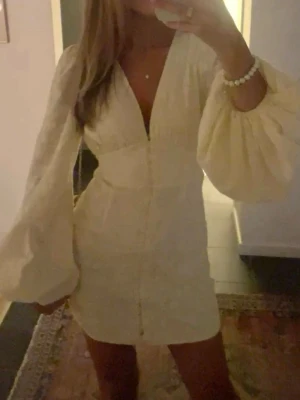 Vit kortklänning med puffärmar - Trendig vit kortklänning med djup v-ringning och långa puffärmar. Klänningen har en figurnära passform och är tillverkad i ett lätt material som passar perfekt till varma dagar. Enkel men elegant design som ger en chic look.