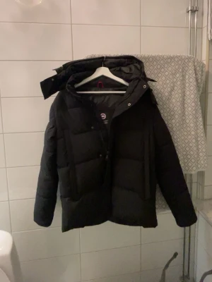 Svart dunjacka från Canada Goose - Säljer en svart dunjacka från Canada Goose med huva och klassisk logga på ärmen. Jackan har en puffig design, dragkedja och tryckknappar framtill samt två stora fickor. Perfekt för kalla vinterdagar och riktigt varm.