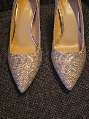 Glittriga pumps från Michael Kors - Snygga pumps från Michael Kors i glittrigt silvermaterial med spetsig tå och smal klack. Skorna har dekorativa stenar över hela ytan och en elegant detalj med metall och sten bak på hälen. Perfekta för att sticka ut och ge extra bling till din outfit.