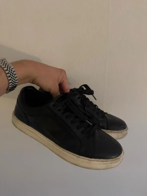 Svarta låga sneakers i läder - Snygga svarta sneakers i läder med vita sulor och klassisk snörning. Skorna har en stilren och minimalistisk design som passar till det mesta. Perfekta för dig som gillar en clean och enkel look.